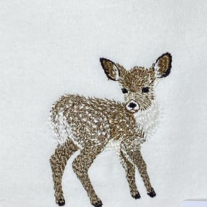May include: An embroidered image of a fawn, featuring brown and white thread detailing. The fawn stands on four legs, with its head turned slightly to the side. The embroidery is on a white background, showcasing the detailed stitching.