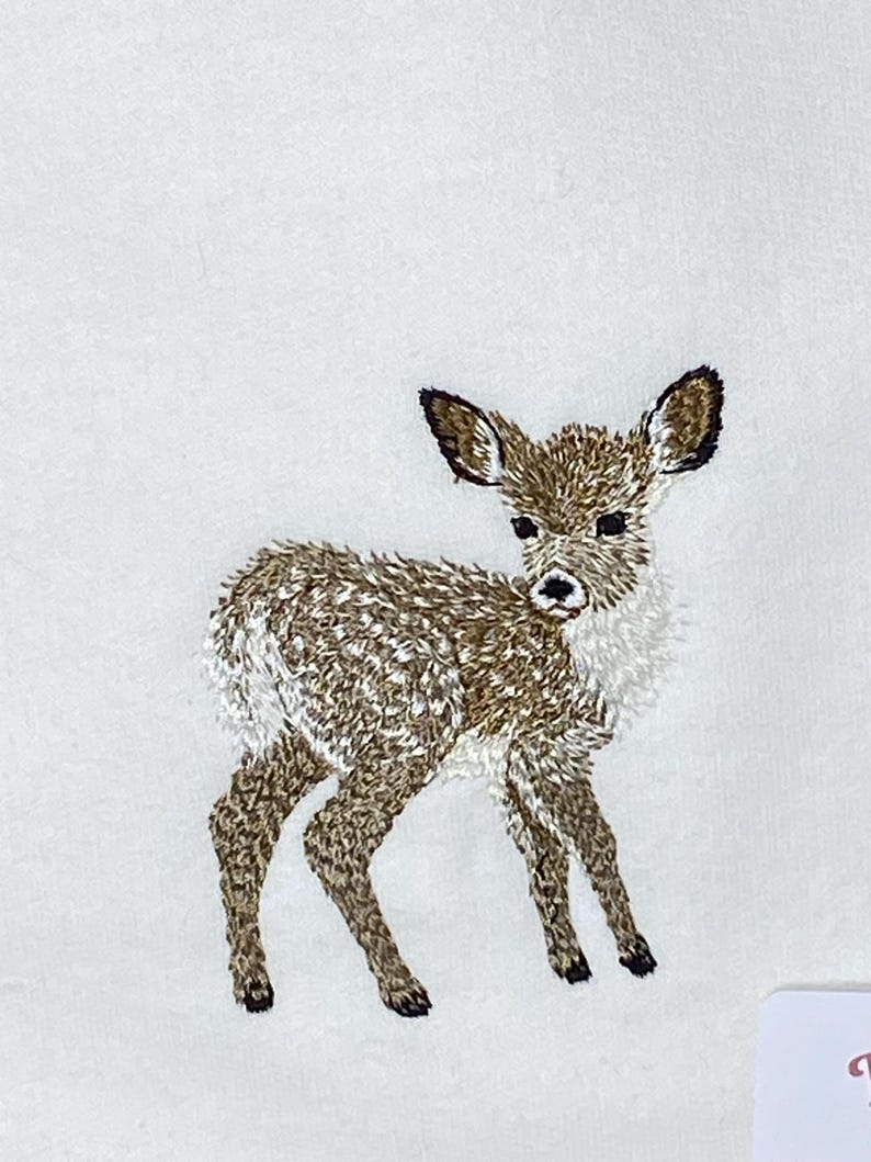 May include: An embroidered image of a fawn, featuring brown and white thread detailing. The fawn stands on four legs, with its head turned slightly to the side. The embroidery is on a white background, showcasing the detailed stitching.