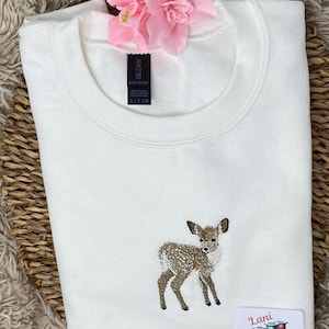 May include: White sweatshirt with an embroidered fawn design. The fawn is brown and white with black accents. A pink flower and a small label with the text "Lani Embroidery" are also visible.