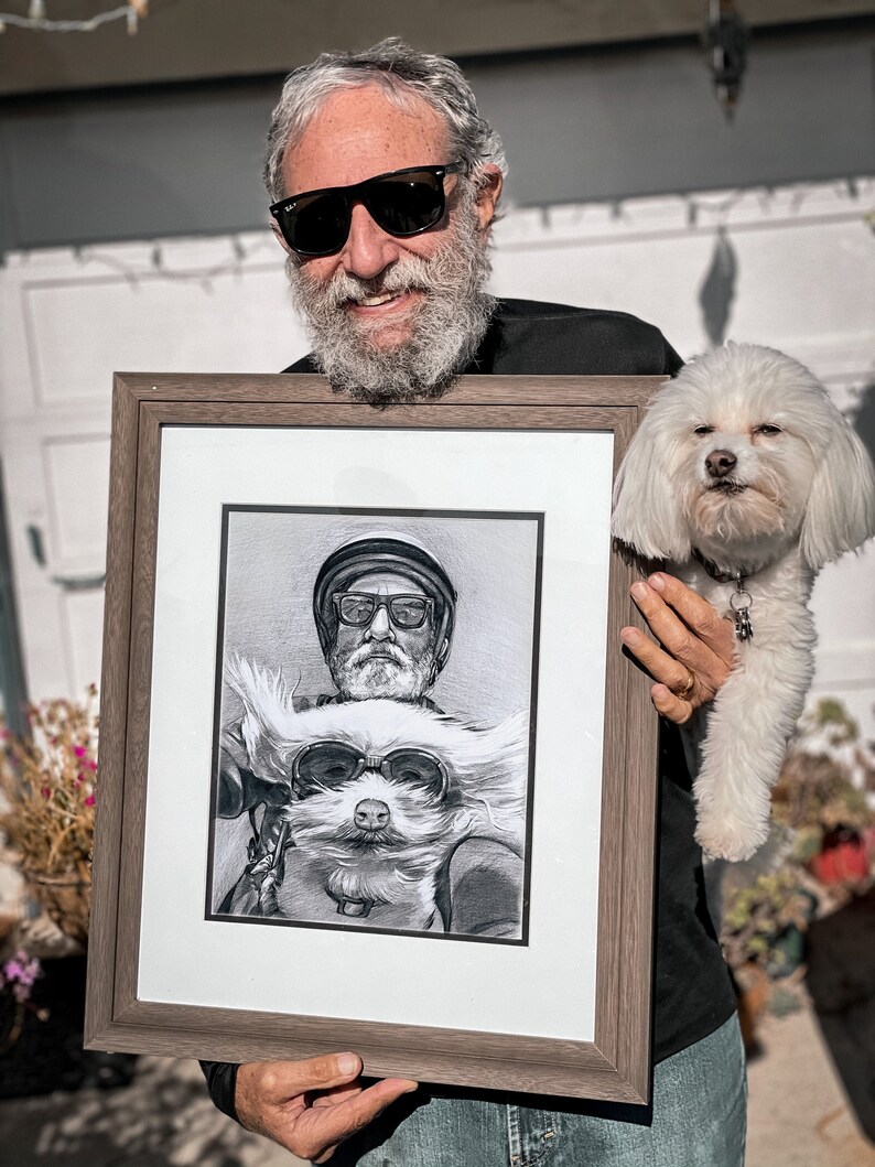 May include: A framed black and white pencil drawing of a man and a dog wearing sunglasses. The man is wearing a hat and the dog is wearing goggles. The drawing is in a brown frame.