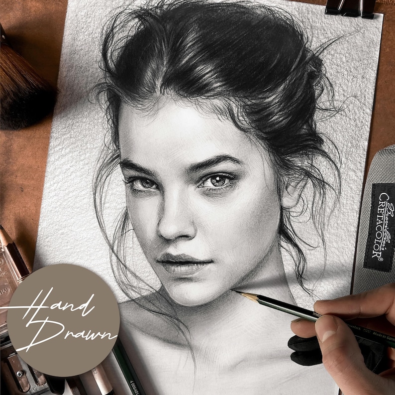May include: A detailed pencil drawing of a woman's face, showcasing realistic shading and fine lines. The artwork is held with a clip, and the words "Hand Drawn" are visible. A makeup brush and pencils are in the background.