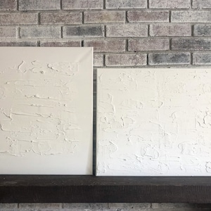 May include: Two white textured canvas paintings with a raised, rough surface. The paintings are displayed on a dark brown shelf against a brick wall.