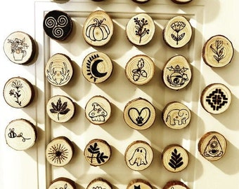 May include: A collection of 27 wooden round ornaments with various designs, including a crescent moon, a pumpkin, a heart, a cat, a ghost, a mushroom, a sun, a flower, a plant, a bee, a mountain, a cauldron, a deer, an acorn, an elephant, a witch, a triple moon symbol, a spiral, a tree, a mandala, and a ghost. The ornaments are all made of natural wood and have a rustic, handmade look.