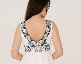 May include: A white sleeveless dress with a floral embroidered neckline and back. The dress has a flowing skirt and is worn by a person. The floral embroidery includes green, blue, and beige tones.