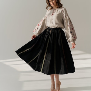 May include: A traditional outfit comprising a beige linen blouse with embroidered sleeves and a black skirt. The blouse has a tie-neck detail, and the skirt features decorative vertical stripes. Suitable for cultural events or everyday wear.