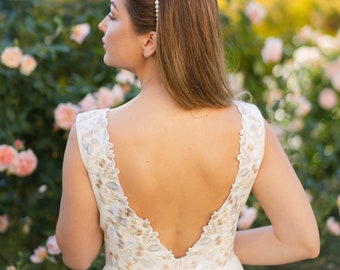 May include: A white wedding dress with a low V-back and floral embroidery on the shoulders. The bride is wearing a pearl headband. The background is a garden with pink roses.