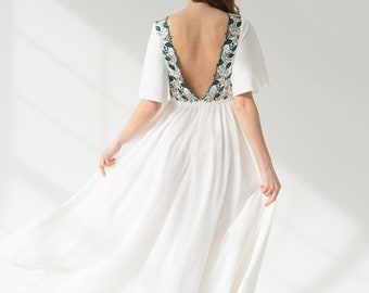 May include: A white maxi dress with a low back and floral embroidery along the neckline. The dress has short sleeves and a flowing skirt.