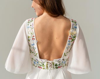 May include: White dress with floral embroidery on the bodice and sleeves. The dress features a low back and a lace-up closure. The embroidery includes blue, red, and green floral designs. The sleeves are wide and flowing.