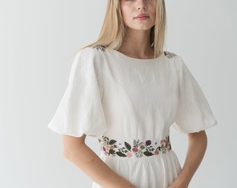 May include: A white linen dress with a floral embroidered waistband. The dress has a round neckline and short, puffed sleeves.