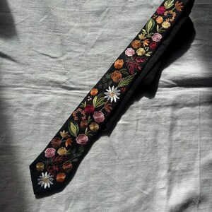 May include: A black necktie featuring intricate floral embroidery along its length. The design includes various flowers in shades of white, pink, yellow, orange, and deep red, accented with green leaves and stems, creating a detailed botanical pattern.