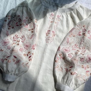 Op de afbeelding: Crème linnen blouse met gedetailleerde bloemenborduurwerk in roze en bruin op de mouwen en halslijn. De blouse heeft een ronde hals en lange, gepofte mouwen, wat een delicate en vrouwelijke uitstraling geeft.