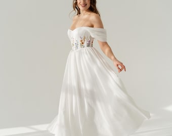 May include: A white off-the-shoulder dress featuring a sweetheart neckline and a bodice adorned with colorful floral embroidery. The garment has a flowing, floor-length skirt and is paired with white strappy sandals, creating a cohesive look.