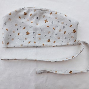Könnte beinhalten: Eine weiße Stoff-Operationsmütze mit einem braunen und weißen Muster aus Blättern, Pilzen und Vögeln. Die Mütze hat eine lange Schleife, die am Hinterkopf gebunden werden kann.