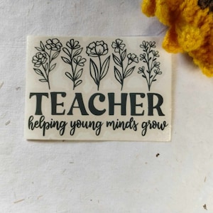 May include: A white sticker with black text and floral illustrations. The text reads "TEACHER helping young minds grow".