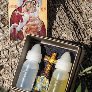 Op de afbeelding: Een icoon van de Maagd Maria en het Christuskind, met tekst, staat boven een doos met twee kleine flesjes en een gouden kruisvormige hanger. De flesjes zijn transparant met witte doppen.