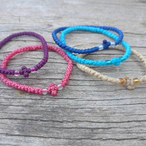 Könnte beinhalten: Eine Sammlung farbenfroher, handgefertigter Armbänder in Lila, Rosa, Blau und Beige. Jedes Armband hat ein geknotetes Design mit dekorativen Akzenten. Die Armbänder sind auf einer Holzoberfläche ausgestellt.
