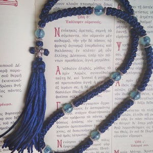 May include: A blue and silver prayer rope with blue and green beads. The rope is laid out on a page of a book with Greek text. The text includes the words "Ωδὴ δ'" and "Ἐκάλυψεν οὐρανούς".