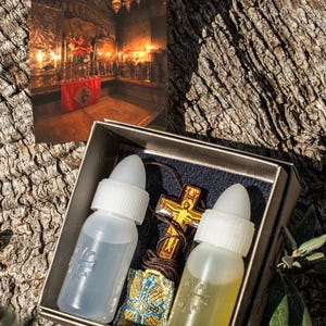Pode incluir: Uma pequena caixa de madeira contendo um colar com uma cruz de madeira marrom, uma pequena bolsa azul e dourada e dois frascos de vidro transparente com tampas brancas. A caixa está etiquetada como "Monastery Blessing".