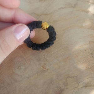 Könnte beinhalten: Ein schwarzer Gebetskranzring mit einem goldfarbenen Kreuz. Der Ring besteht aus geknotetem schwarzem Kordel. Das Kreuz ist auf dem Ring zentriert. Der Hintergrund ist eine hellbraune Holzoberfläche.