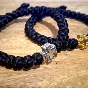 Könnte beinhalten: Drei schwarze geflochtene Armbänder mit silbernen und goldenen Kreuzanhängern. Die Armbänder sind geknüpft und haben ein schlichtes Design.
