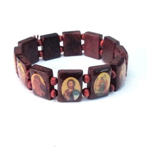 Può includere: Un braccialetto di legno con immagini di figure religiose. Il braccialetto è in legno marrone scuro e ha un filo rosso che collega le perline.