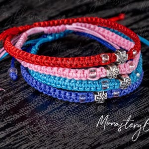 Op de afbeelding: Vier gevlochten armbanden met zilveren kruisbedels. De armbanden zijn rood, roze, blauw en donkerblauw.
