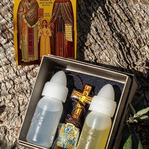 Op de afbeelding: Een klein, bruin doosje met een gouden rand bevat twee heldere glazen flesjes met witte dopjes, een klein blauw en gouden zakje en een bruine houten kruis ketting. Het doosje ligt op een boomstam. De tekst "Monastery Blessing" staat op de onderkant van het doosje.