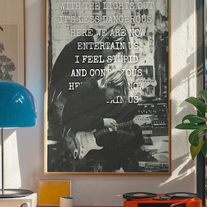 May include: A framed black and white wall art print featuring a musician playing an electric guitar with the lyrics: WITH THE LIGHTS OUT IT'S LESS DANGEROUS HERE WE ARE NOW ENTERTAIN US I FEEL STUPID AND CONTAGIOUS HERE WE ARE NOW ENTERTAIN US.
