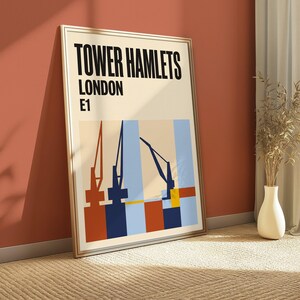 May include: A framed wall art print featuring the text TOWER HAMLETS LONDON E1 in bold black letters. The graphic design displays stylized industrial cranes in dark blue and red against a light cream background with geometric color blocks.