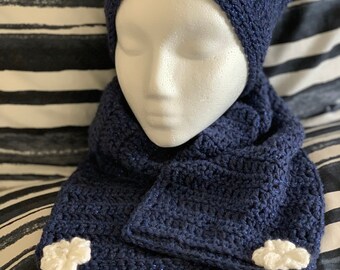 May include: A navy blue crochet hat and scarf set. The hat is adorned with three white flower appliqués. The scarf has a matching white flower appliqué on one end. The set is displayed on a mannequin head.