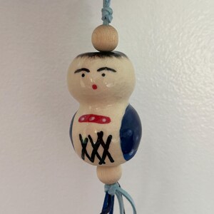 May include: A hand-painted ceramic doll ornament with a light blue string. The doll has a cream-colored face with black eyes and a red mouth. The doll's body is white with blue and red accents and black markings.
