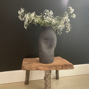 May include: A black ceramic vase with a textured surface holds a bouquet of white baby's breath flowers. The vase is sitting on a rustic wooden table with three legs.