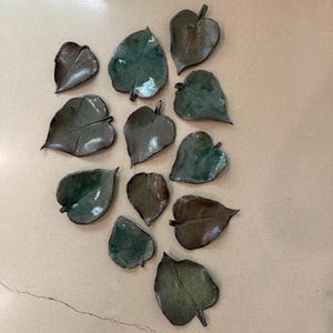 May include: A collection of ceramic leaf-shaped dishes in varying shades of green and brown. The leaves have a glossy finish and are arranged on a neutral-toned surface. The dishes are likely for decorative or functional use.