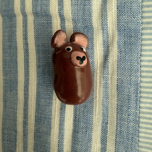 May include: A small, smooth, dark brown ceramic figurine shaped like a bean with two rounded ears. It features a light pink snout with a black nose, two white eyes with black pupils, and a glossy finish that reflects light across its rounded surface.