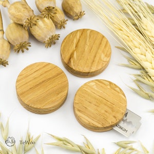 Könnte beinhalten: Drei runde Holzobjekte mit hellbraunem, gestreiftem Maserungsmuster. Sie sind auf einer weißen Oberfläche mit getrocknetem Weizen, Mohnkapseln und einer Rasierklinge angeordnet. Die Objekte scheinen Untersetzer oder kleine Dekorationsartikel zu sein.