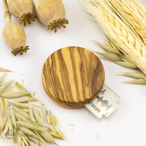Könnte beinhalten: Ein runder Rasierklingenhalter aus Holz mit einem Holzmaserungsmuster. Eine silberne Rasierklinge ist in den Halter eingesetzt. Das Bild enthält auch getrockneten Weizen, Mohnkapseln und Hafergras.