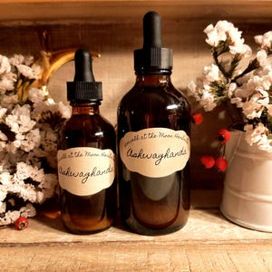 Puede incluir: Dos botellas de vidrio marr&oacute;n con cuentagotas negros y etiquetas que dicen "Well at the Moon Herbals Ashwagandha". Las botellas est&aacute;n sobre una superficie de madera, rodeadas de flores blancas y bayas rojas.