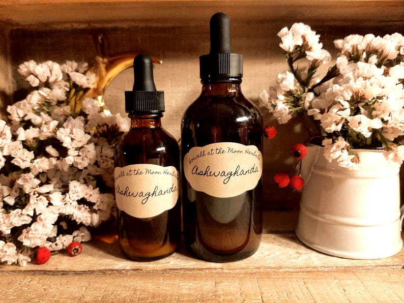 Puede incluir: Dos botellas de vidrio marr&oacute;n con cuentagotas negros y etiquetas que dicen "Well at the Moon Herbals Ashwagandha". Las botellas est&aacute;n sobre una superficie de madera, rodeadas de flores blancas y bayas rojas.