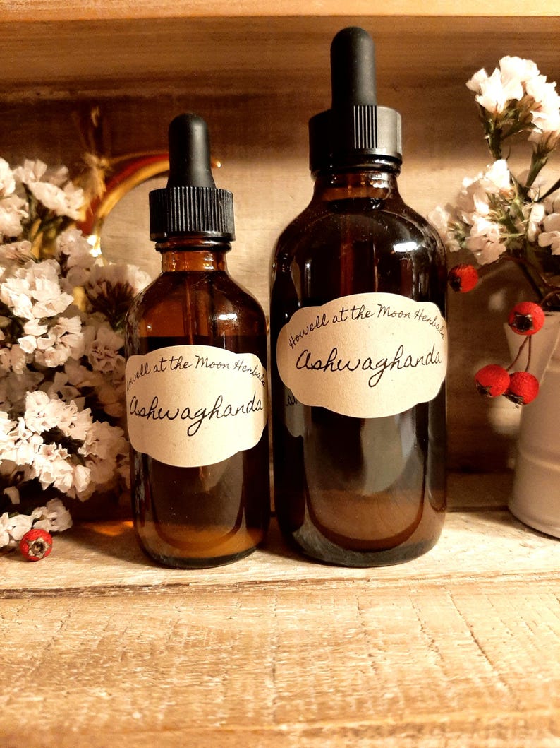 May include: Two amber glass bottles with black droppers. Each bottle has a cream-colored label with the text "Howell at the Moon Herbals" and "Ashwaghanda" in handwritten script. The bottles are displayed on a wooden surface, with white flowers and red berries in the background.