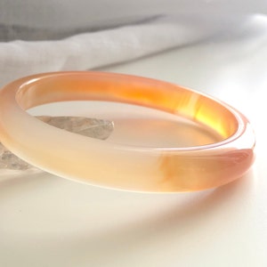Puede incluir: Un brazalete de ágata de color naranja claro y blanco. El brazalete es liso y pulido.