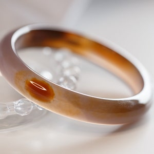 Puede incluir: Un brazalete de ágata de color marrón claro y blanco con una mancha marrón rojiza en el centro.