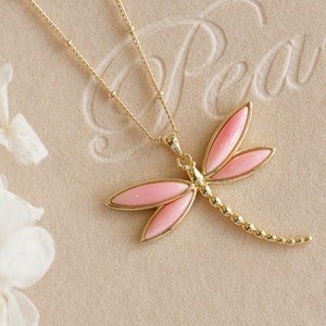 May include: A gold necklace with a pink dragonfly pendant. The dragonfly has a beaded tail.