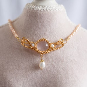 Puede incluir: Un collar de oro con un colgante de cuarzo rosa y detalles de perlas. El collar est&aacute; en un busto de maniqu&iacute; blanco.
