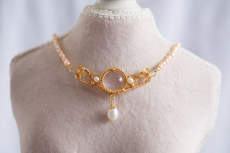 Puede incluir: Un collar de oro con un colgante de cuarzo rosa y detalles de perlas. El collar est&aacute; en un busto de maniqu&iacute; blanco.