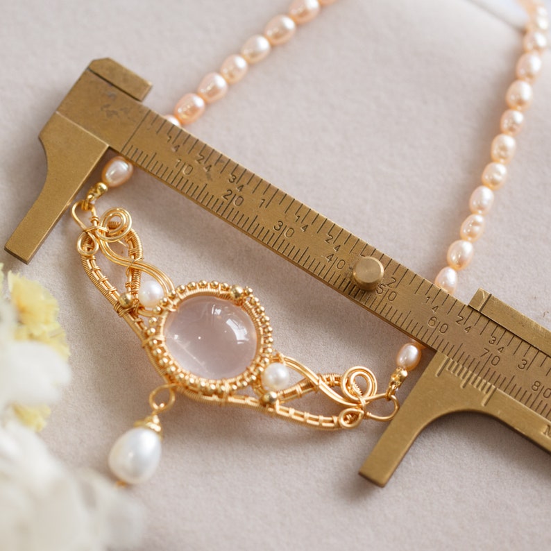 Puede incluir: Un collar con un colgante de alambre dorado, una piedra de cuarzo rosa y detalles de perlas. El collar se mide con una regla, mostrando que mide aproximadamente 5 cm de largo.