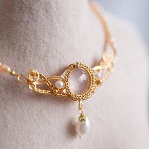 Puede incluir: Un collar de oro con un delicado dise&ntilde;o de alambre presenta un gran colgante de piedra preciosa redonda y transparente con una perla m&aacute;s peque&ntilde;a que cuelga debajo. El collar est&aacute; acentuado con peque&ntilde;as perlas y cuentas verdes.