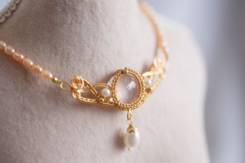 Puede incluir: Un collar de oro con un delicado dise&ntilde;o de alambre presenta un gran colgante de piedra preciosa redonda y transparente con una perla m&aacute;s peque&ntilde;a que cuelga debajo. El collar est&aacute; acentuado con peque&ntilde;as perlas y cuentas verdes.