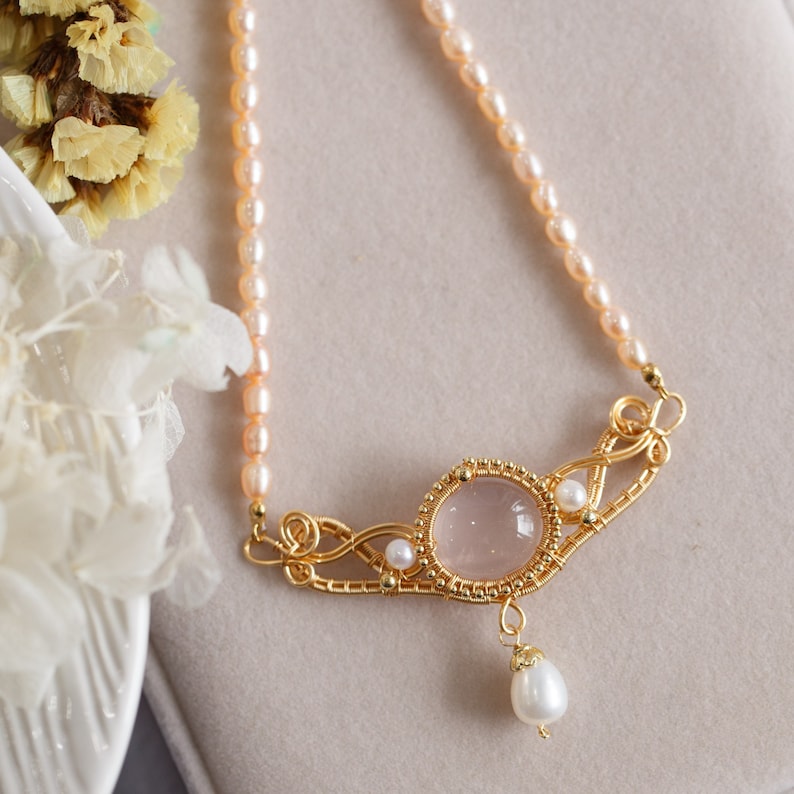 Puede incluir: Un delicado collar de oro con un colgante de piedra rosa y una gota de perla. El collar est&aacute; hecho con un dise&ntilde;o de alambre delicado y presenta una hebra de peque&ntilde;as perlas rosas.