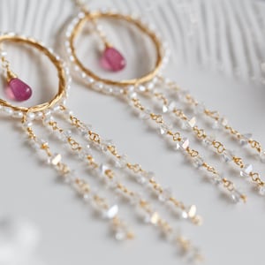 May include: Gold hoop earrings with a pink gemstone dangle and a long chain of clear crystals.