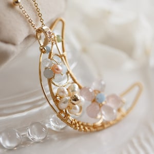 May include: A gold-colored crescent moon pendant necklace. The pendant is adorned with small, delicate flowers made of pearls and pink and blue gemstones. The necklace has a delicate chain.
